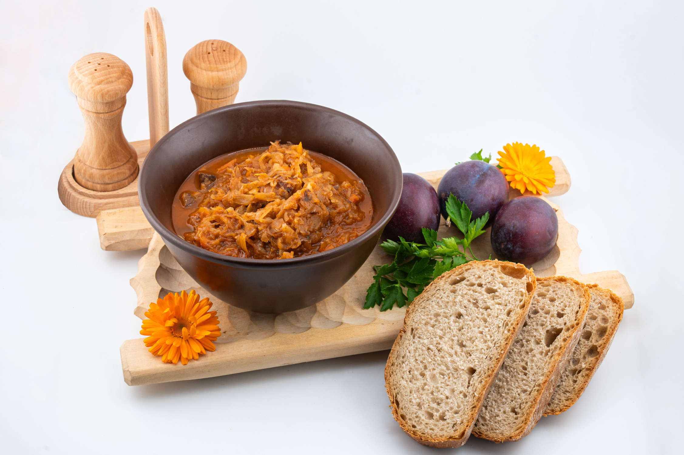 Propozycja podania bigosu z dzika. Danie znajduje się w brązowej misce podane na drewnianej desce z pieczywem oraz dekoracją.